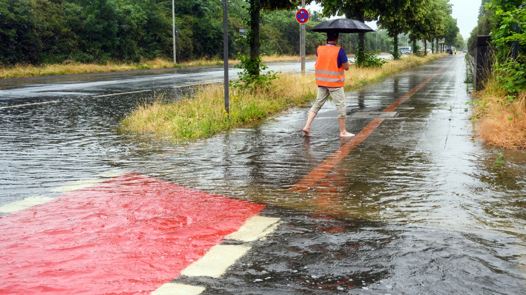 Foto: Bei heftigem Regen watet ein Passant barfüßig mit Regenschirm und Warnweste über eine überschwemmte Straßenkreuzung
